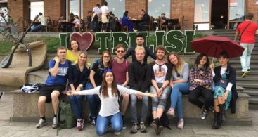 part of the group, cheerful as always, sits in front of the inscription "I love Tbilisi". Two girls hold an umbrella wide open above themselves.