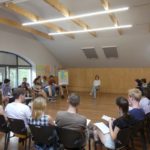 the whole group sits in the semi-circle facing the trainer. There is a flipchart on the left which says "Migration - Challenge for the World"