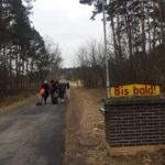 По дорозі в лісі йдуть учасники проекту, видно лише їхні спини. На передньому плані табличка німецькою "Bis bald!", що означає "Скоро побачимось!".