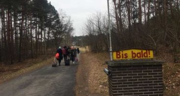 По дорозі в лісі йдуть учасники проекту, видно лише їхні спини. На передньому плані табличка німецькою "Bis bald!", що означає "Скоро побачимось!".