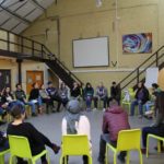 Учасники сидять в колі, їхня увага звернена на тренера. Позаду стоїть фліпчарт, а також на стіні висить мультимедійний екран.