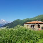 Група людей в поході йдуть в один ряд один за одним. На фоні видніється хатинка, зелені гори та гори вкриті снігом.