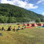 Учасники сидять біля гірської річки, схрестивши ноги, їхнє взуття лежить поруч. Долоні учасників лежать на колінах внутрішньою частиною догори. Зліва стоять сині та червоні палатки.