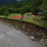 Фото поділене на дві частини: зліва тече гірська річка, справа розташований кемпінг, оточений зеленими деревами, маленькі групки людей стоять біля палаток.