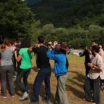 Учасники рухаються в колі, виконуючи вправи руками. На фоні видніється багато зелених дерев.