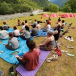 Учасники сидять біля гірської річки на карематах, схрестивши ноги, їхнє взуття лежить поруч. Долоні учасників лежать на колінах внутрішньою частиною догори. На фоні стоять сині та червоні палатки.