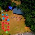 Фото зняте з висоти. Зліва розташований кемпінг, де є багато червоних, синіх палаток та одні жовта. Справа розташований великий тент, біля нього учасники виклали напис “Breathe in!”( “Вдихни!”) своїми тілами. Територія оточена багатьма деревами.