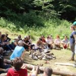 Учасники сидять на землі у півколі, одна людина сидить на колоді. Двоє людей стоять перед ними та презентують щось біля фліпчарту.