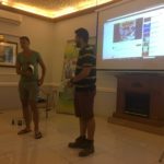 Двоє людей презентують щось в кімнаті з підлогою вкритою плиткою. Позаду них є екран, банер та камін. Справа на стіні висить картина та кондиціонер над нею.