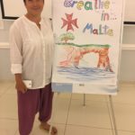 Жінка стоїть біля фліпчарту, де написано “Breathe in Malta” та намальовані море та скеля. Вона вдягнути у вільні штани та сорочку. В правій руці тримає телефон.