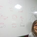 Two schoolgirls are next to the whiteboard. One of them is drawing, the other one is smiling at the camera.