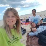 Three girls and one boy are on an almost empty beach. One of the girls is taking a selfie, others are lying on the towel, the boy is sitting next to them.