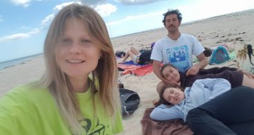 Three girls and one boy are on an almost empty beach. One of the girls is taking a selfie, others are lying on the towel, the boy is sitting next to them.