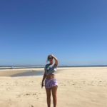 The girl, wearing shorts and a T-shirt, is standing on an almost empty beach and holding her hand at her head.
