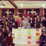 A group photo of international team. Five people in the middle are holding a bid flipchart paper with colorful inscriptions and papers on it.