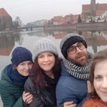 Three girls and one boy, wearing jackets, hats, and scarfs, are taking selfie next to a lake. There is also a view of old town on the background